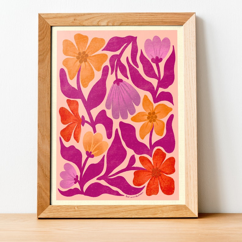 May include: A framed print with a pink background and a floral pattern in shades of orange, pink, and red. The flowers are stylized and have a retro feel. The print is signed "but we thrive" in the bottom right corner.