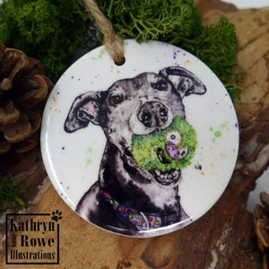 May include: A white ceramic ornament featuring a black and white illustration of a dog holding a green and purple toy. The background is white with green, purple, and yellow splatters.