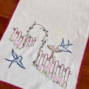 May include: A rectangular off-white table runner featuring embroidered blue swallows, a garden scene with a pink picket fence, green vines, and a birdbath with a yellow water spray. The edges are finished with a decorative dark pink crochet border.