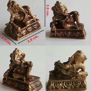 Może przedstawiać: Mała, brązowa statua Ganeshy o wymiarach 2,0 cm długości, 1,2 cm szerokości i 2,0 cm wysokości. B&oacute;stwo przedstawione jest w pozycji leżącej na ozdobnej podstawie. Posąg ma skomplikowane detale i czerwonobrązowy odcień.