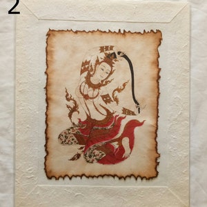 May include: A framed print of a traditional Thai painting depicting a woman in a red and gold dress, holding a black ribbon. The painting is on aged paper with a textured border.
