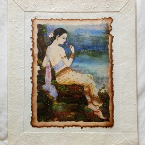 May include: A framed watercolor painting of a woman in a traditional Thai dress sitting on a rock by a body of water. She is holding a flower in her hand and looking down at it. The painting is set against a backdrop of a blue sky and clouds.