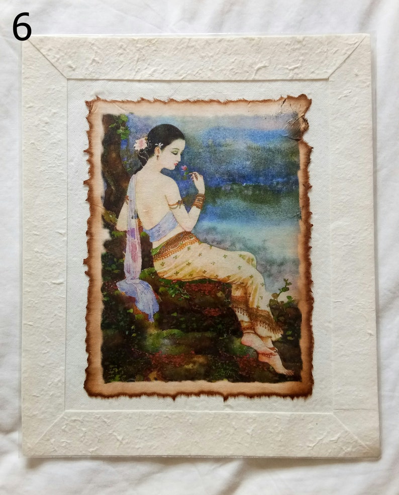 May include: A framed watercolor painting of a woman in a traditional Thai dress sitting on a rock by a body of water. She is holding a flower in her hand and looking down at it. The painting is set against a backdrop of a blue sky and clouds.