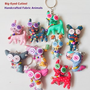 May include: A collection of fabric animal keychains labeled Big-Eyed Cuties! Handcrafted Fabric Animals. Each keychain features a small stuffed animal made from patterned fabric with large button eyes, a beaded collar, and a metal ring attachment.