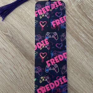 May include: A black bookmark with a purple tassel. The bookmark features a repeating pattern of pink video game controllers and hearts with the name "Freddie" written in pink letters.