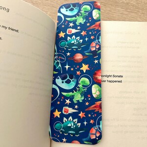 May include: A blue bookmark with a space-themed dinosaur pattern. The design includes green dinosaurs, planets, stars, and rockets. The bookmark is placed in a book with text visible on the pages.