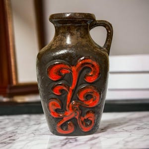 Peut inclure: Un vase pichet en céramique brun foncé avec une finition brillante et un motif en relief proéminent de couleur orange vif. Le récipient possède une anse sur le côté droit, un col large et un vernis texturé et moucheté sur tout le corps.