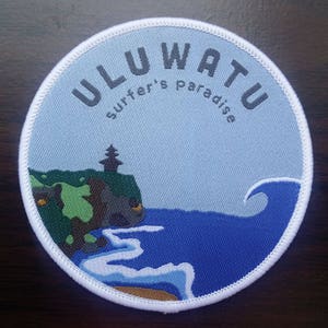 Könnte beinhalten: Ein weißer, runder Patch mit blauen und weißen Wellen, einer grünen Klippe und einem kleinen Tempel. Der Text "ULUWATU surfer's paradise" ist in Schwarz auf dem Patch gedruckt.