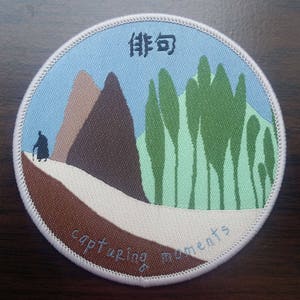 Pode incluir: Remendo redondo com um cenário de paisagem bordado. Apresenta montanhas, árvores e uma pessoa com uma bengala. O texto "capturing moments" é costurado na parte inferior, com caracteres japoneses no topo.