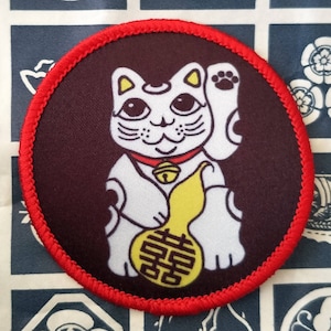 Op de afbeelding: Een witte Maneki-Neko, of gelukkat, met een gele kalebas en een rode halsband met een bel. De kat zit op een donkerbruine achtergrond met een rode gestikte rand.