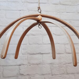 May include: A wooden baby mobile frame with four arches, a wooden connector, and a metal hook for hanging. The frame is made of natural wood and has a light brown color.