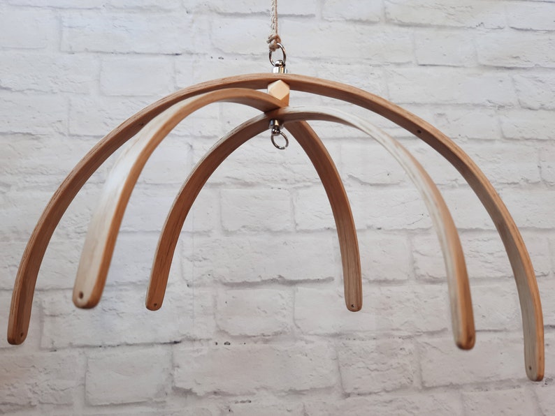 May include: A wooden baby mobile frame with four arches, a wooden connector, and a metal hook for hanging. The frame is made of natural wood and has a light brown color.