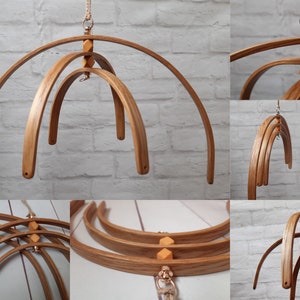 May include: A hanging mobile constructed from multiple curved wooden arches arranged in a nested, circular formation. The structure features light brown wood grain, small geometric wooden beads at the connection points, and silver metal hardware.