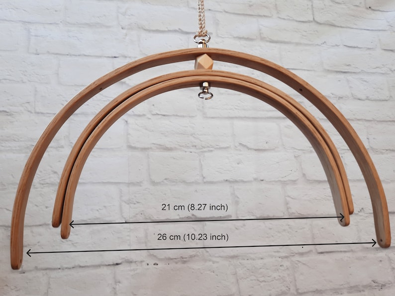May include: Two wooden hoops, one 21 inches in diameter and the other 26 inches in diameter, are hanging from a white brick wall. The hoops are connected by a metal ring and a wooden bead.
