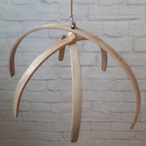 May include: A wooden baby mobile frame with five curved arms, made of natural wood. The frame is suspended from a metal hook.