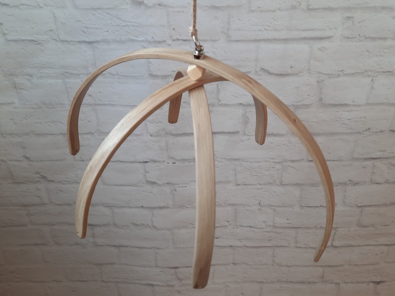 May include: A wooden baby mobile frame with five curved arms, made of natural wood. The frame is suspended from a metal hook.