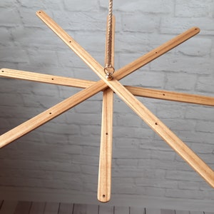 Puede incluir: Móvil de madera con ocho brazos rectangulares de color marrón claro que irradian desde un punto central. Cada brazo tiene tres pequeños agujeros y está suspendido por una cuerda. Diseñado para decoración.