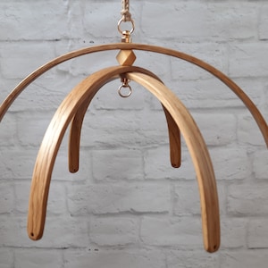 May include: A wooden mobile frame with three curved wooden arches. The frame is made of natural wood and has a gold metal ring at the top.