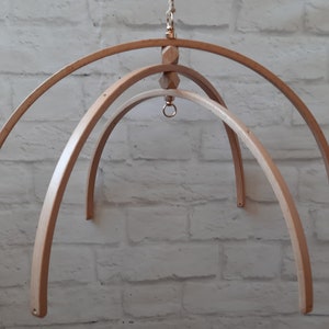 Puede incluir: Un colgador de móvil de madera con tres aros circulares, una cuenta de madera y un anillo de metal. El colgador está suspendido de una pared de ladrillo blanco.