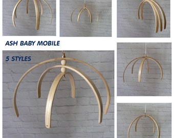 Puede incluir: Se muestran varios modelos de móviles para bebés de madera de fresno. Cada móvil presenta arcos de madera curvados suspendidos de una cuerda, con un acabado de madera natural. Se muestra el texto "ASH BABY MOBILE" y "5 STYLES".