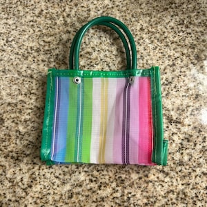 May include: A small, colorful tote bag with a rainbow striped design. The bag features a green trim and handles. The bag is made of a mesh-like material, and the handles are a solid green color.
