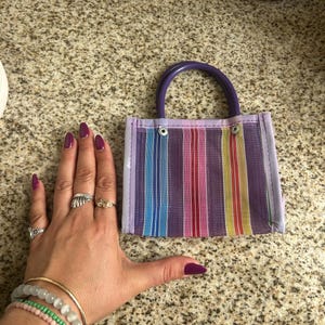 May include: A small, rectangular tote bag with a purple handle and a striped pattern in rainbow colors. The bag is made of a woven material and has a light purple border. The bag is approximately 4 inches wide.