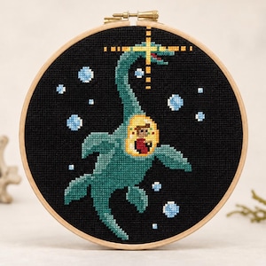 May include: A circular wooden embroidery hoop containing a cross-stitch design on black fabric. The design features a teal sea creature with a yellow porthole on its body, revealing a small figure inside, surrounded by light blue bubbles and a yellow star.