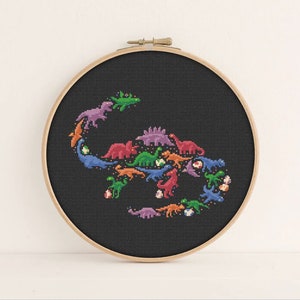 May include: A black embroidery hoop featuring a colourful cross-stitch design of dinosaurs arranged in a circle. The dinosaurs are in a variety of colours, including green, blue, purple, orange and red.