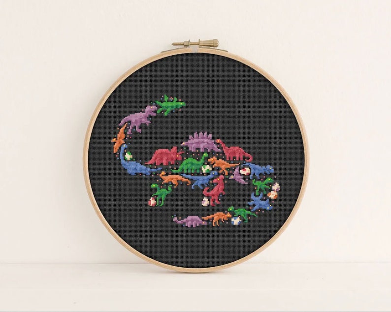 May include: A black embroidery hoop featuring a colourful cross-stitch design of dinosaurs arranged in a circle. The dinosaurs are in a variety of colours, including green, blue, purple, orange and red.