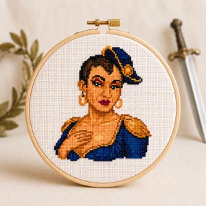 May include: A circular wooden embroidery hoop containing a cross-stitch portrait of a person with short dark hair, wearing a blue military-style jacket with gold epaulets, a matching blue cap with a gold emblem, and large gold hoop earrings.