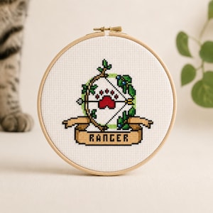May include: A circular wooden embroidery hoop containing a cross-stitch design on white fabric. The design features a bow and arrow intertwined with green vines, a red paw print in the center, and a tan banner below with the text RANGER.
