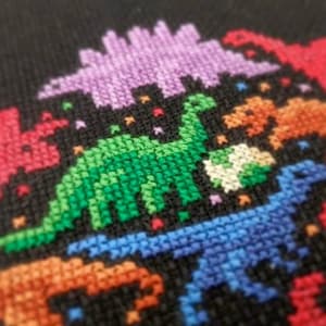 May include: A close-up of a colourful cross-stitch design featuring a variety of dinosaurs in different colours, including green, blue, purple, orange, and red. The dinosaurs are stitched on a black fabric background.
