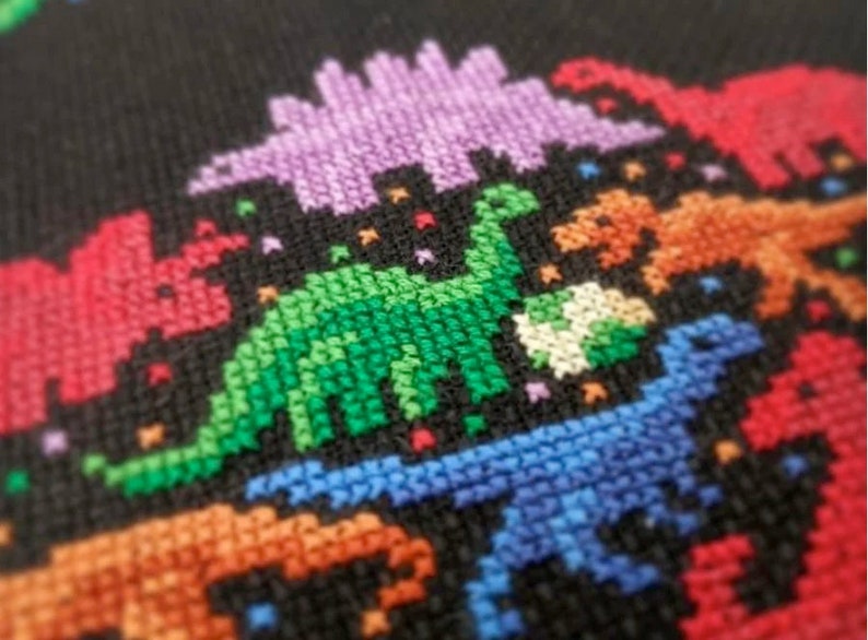 May include: A close-up of a colourful cross-stitch design featuring a variety of dinosaurs in different colours, including green, blue, purple, orange, and red. The dinosaurs are stitched on a black fabric background.