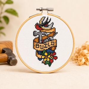 Può includere: Ricamo a punto croce in un telaio di legno. Il disegno presenta un uccello, un martello e altri strumenti con uno striscione che recita "BUILD". La tavolozza dei colori comprende rosso, giallo, blu e marrone.