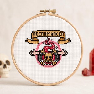 May include: A circular wooden embroidery hoop containing a cross-stitch design on white fabric. The design features a skull with crossed bones, a red swirling smoke element, and a tan banner with the black text NECROMANCER centered above.