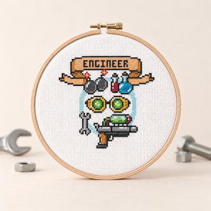 May include: A cross-stitch embroidery in a wooden hoop features an engineer-themed design. The design includes the word "ENGINEER" on a banner, bombs, goggles, lab equipment, and a ray gun. The color palette includes shades of blue, green, and brown.