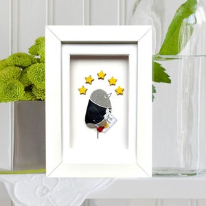 May include: A white framed picture with a gray and black bird with a red beak and a yellow star above it. The bird is holding a small white paper with the words "You are loved" written on it.