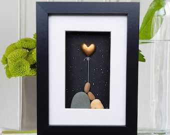May include: A framed pebble art piece depicting a heart-shaped balloon floating above a stack of stones against a black background with white stars. The frame is black with a white mat.