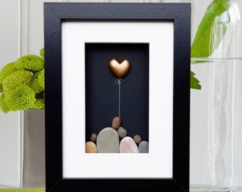 May include: A framed artwork featuring a gold heart-shaped balloon floating above a stack of painted stones. The artwork is set against a black background and framed in black with a white mat.