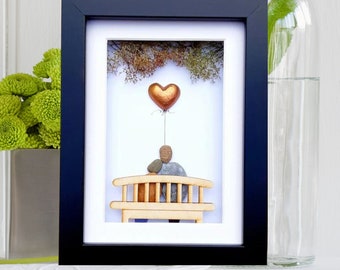 May include: A framed artwork featuring a couple made of stones sitting on a wooden bench. A gold heart balloon floats above them. The artwork is set against a white background.