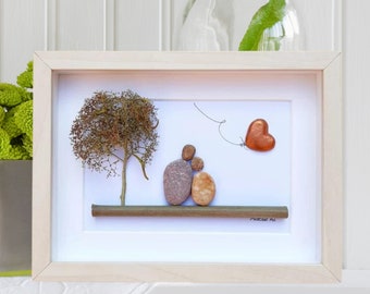 May include: A framed pebble art piece depicting a couple sitting on a rock with a heart-shaped balloon floating above them. The couple is made of small, smooth stones in shades of brown and gray. The background is a white canvas with a small tree in the corner. The frame is a light wood color.