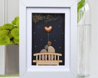 May include: A framed pebble art piece depicting a couple sitting on a bench under a starry night sky. A heart-shaped balloon is floating above them. The artwork is set against a black background and framed in white.