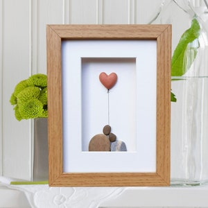 May include: A framed pebble art piece depicting two figures holding a heart-shaped balloon. The figures are made of brown and gray stones, and the balloon is a light pink color. The artwork is set against a white background.