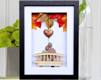 May include: A framed artwork featuring a couple sitting on a wooden bench with a heart-shaped balloon floating above them. The background is decorated with fall leaves in shades of red, orange, and yellow.