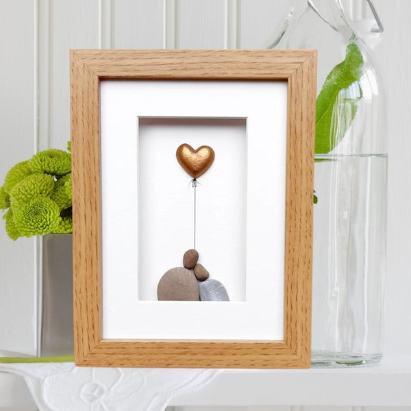May include: A framed artwork featuring a gold heart balloon and two pebble figures. The artwork is in a light wood frame with a white mount.