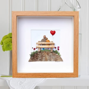 May include: A framed pebble art piece depicting a couple sitting on a bench with a red heart balloon. The couple is made of small stones and the bench is made of twigs. The background is a piece of birch bark with green moss and red flowers.