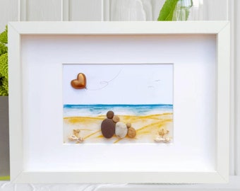 May include: A framed pebble art piece depicting a couple sitting on a beach with a heart-shaped balloon floating above them. The background is a watercolor painting of a beach scene with blue water and yellow sand.