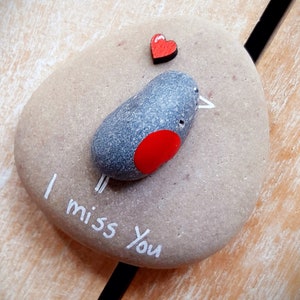 Puede incluir: Una piedra pintada de gris con un p&aacute;jaro pintado de rojo y las palabras "I miss you" escritas en pintura blanca. Un peque&ntilde;o coraz&oacute;n de madera rojo est&aacute; sobre el p&aacute;jaro.