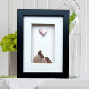 Peut inclure: Une œuvre d'art en galets encadrée représentant une famille de trois avec un ballon en forme de cœur rose. La famille est faite de pierres brunes et grises, et le cœur est fait d'une pierre rose et blanche.