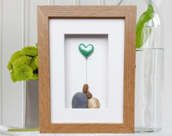 May include: A framed pebble art piece with a green heart-shaped balloon floating above three gray and brown pebbles.
